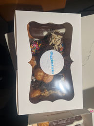 White bakery box with clear window revealing assorted chocolate-coated eclairs, cream puffs and sprinkle-topped doughnuts garnished with almond slivers.