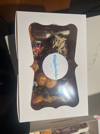 White bakery box with clear window revealing assorted chocolate-coated eclairs, cream puffs and sprinkle-topped doughnuts garnished with almond slivers.