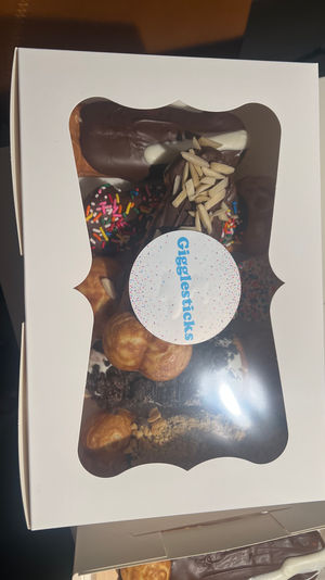 White bakery box with clear window revealing assorted chocolate-coated eclairs, cream puffs and sprinkle-topped doughnuts garnished with almond slivers.