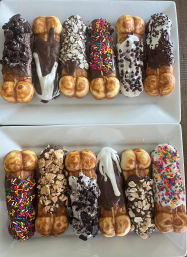 Assorted chocolate-dipped bubble waffle sticks topped with sprinkles, crushed cookies, nuts and white chocolate drizzles arranged on two white rectangular dessert plates