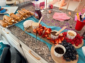 Party dessert spread on a granite kitchen counter with waffle pops on sticks, a wooden board of sliced strawberries, blueberries and raspberries, cups of chocolate dip, whipped cream and crumble toppings, syrup bottle, turquoise runner, pink napkins and confetti