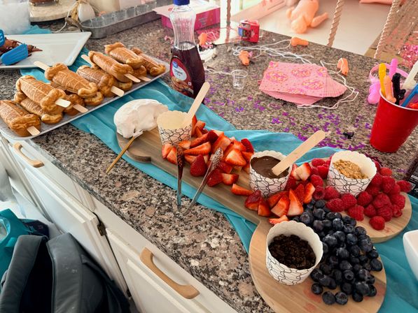 Party dessert spread on a granite kitchen counter with waffle pops on sticks, a wooden board of sliced strawberries, blueberries and raspberries, cups of chocolate dip, whipped cream and crumble toppings, syrup bottle, turquoise runner, pink napkins and confetti
