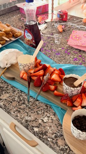 Party dessert spread on a granite kitchen counter with waffle pops on sticks, a wooden board of sliced strawberries, blueberries and raspberries, cups of chocolate dip, whipped cream and crumble toppings, syrup bottle, turquoise runner, pink napkins and confetti