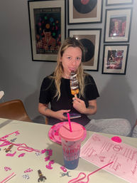 Person sitting at a dining table indoors, playfully licking a chocolate‑drizzled dessert on a stick; pink party decorations, paper plate and lidded cup on the table, framed vinyl record art on the wall behind.
