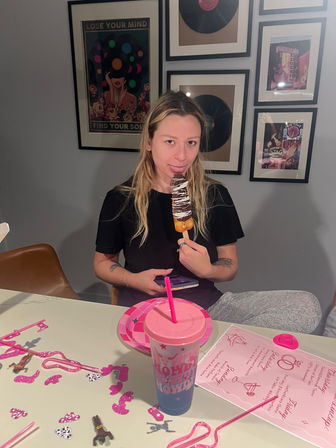 Person sitting at a dining table indoors, playfully licking a chocolate‑drizzled dessert on a stick; pink party decorations, paper plate and lidded cup on the table, framed vinyl record art on the wall behind.