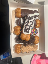 Takeout box of assorted cream-puff eclairs and dipped pastries topped with chocolate, white icing, mini chocolate chips, rainbow sprinkles and cookie crumbs, with a wooden paddle.