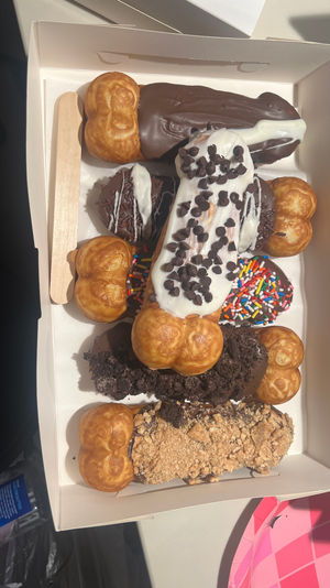 Takeout box of assorted cream-puff eclairs and dipped pastries topped with chocolate, white icing, mini chocolate chips, rainbow sprinkles and cookie crumbs, with a wooden paddle.