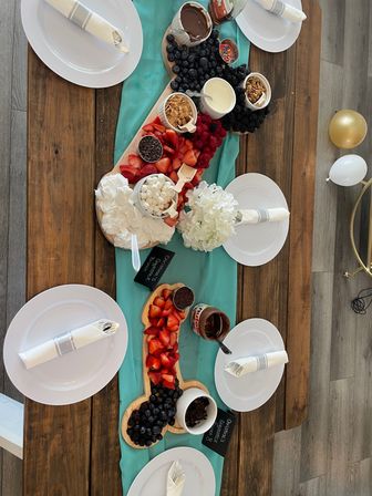 Top-down view of a playful novelty wooden dessert grazing board on a teal runner, loaded with strawberries, blueberries, raspberries, blackberries, marshmallows, whipped cream, chocolate chips, Nutella and nuts, surrounded by white plates with rolled napkins and gold and white balloons.