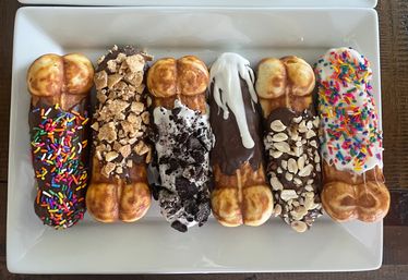 Overhead shot of six dipped waffle sticks on a white rectangular plate, chocolate- and vanilla-coated waffles topped with rainbow sprinkles, crushed cookies, cereal clusters and chopped peanuts — colorful dessert platter