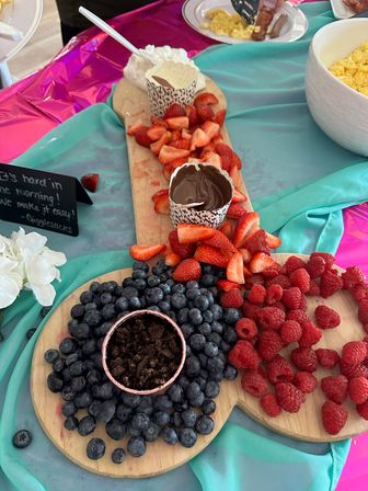 Bright brunch fruit board with sliced strawberries, blueberries, raspberries and chocolate dip in paper cups on wooden serving boards over teal and pink tablecloth.