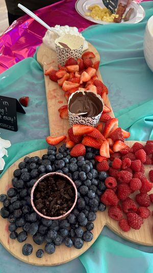 Bright brunch fruit board with sliced strawberries, blueberries, raspberries and chocolate dip in paper cups on wooden serving boards over teal and pink tablecloth.