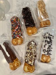 Six individually wrapped chocolate-dipped bubble waffle sticks on a marble countertop, each topped with colorful sprinkles, crushed cookies, graham crumbs, white drizzle, chopped nuts or mini chocolate chips — fun gourmet dessert treats.