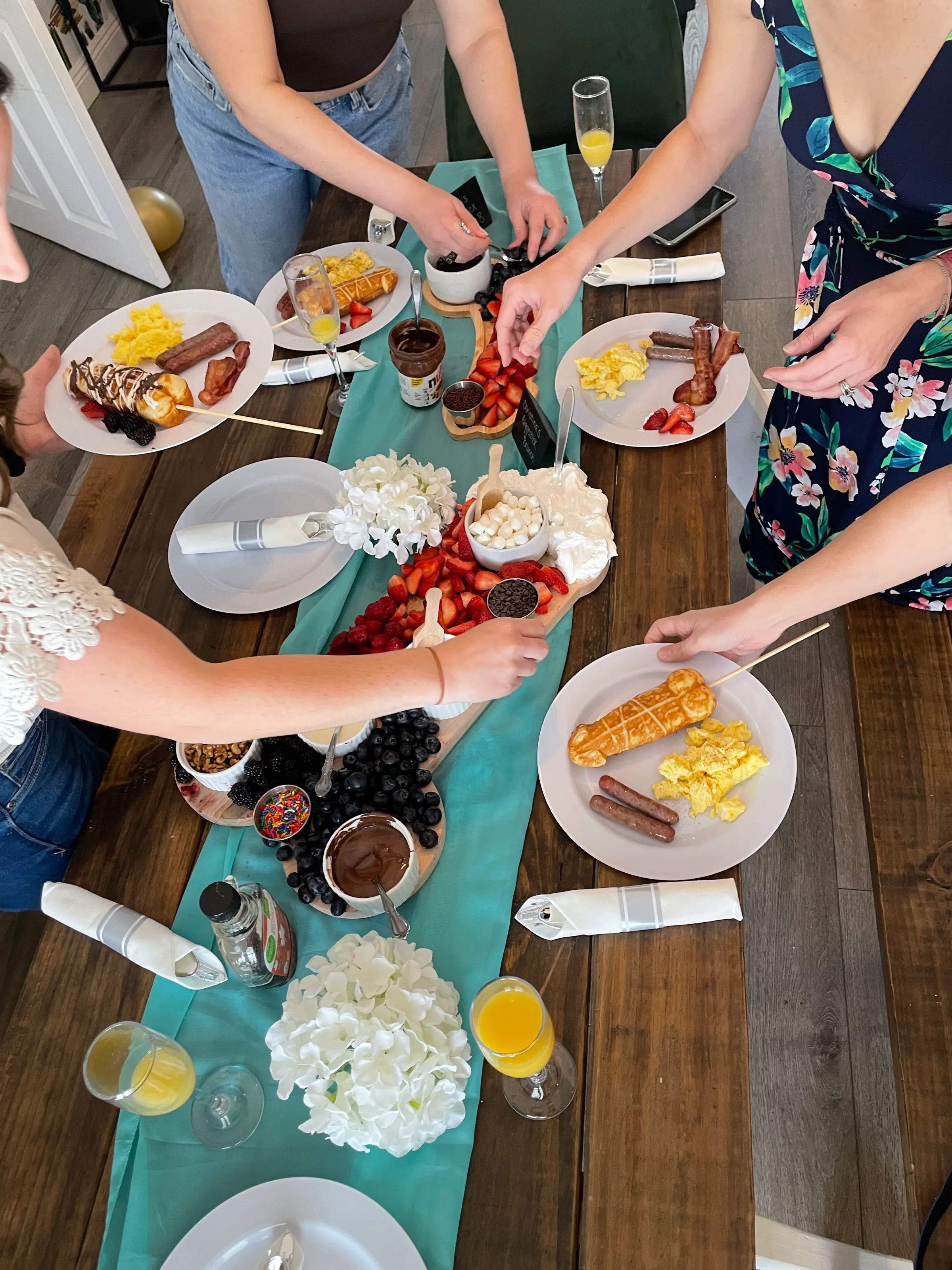 Overhead view of an indoor brunch spread with a teal table runner, wooden boards of strawberries, raspberries, blueberries, marshmallows, chocolate and Nutella, plates of scrambled eggs, sausage and bacon, waffle-on-a-stick, mimosas and guests reaching for fruit.