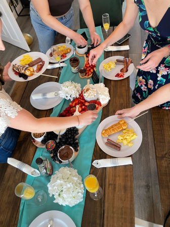 Overhead view of an indoor brunch spread with a teal table runner, wooden boards of strawberries, raspberries, blueberries, marshmallows, chocolate and Nutella, plates of scrambled eggs, sausage and bacon, waffle-on-a-stick, mimosas and guests reaching for fruit.