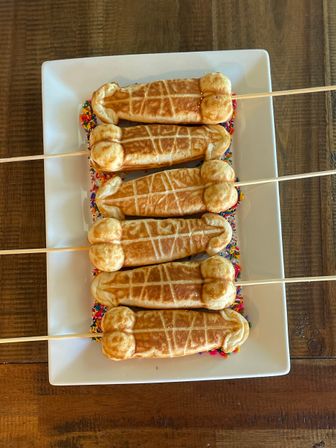 Novelty phallic-shaped waffle pops on wooden skewers with rainbow sprinkles arranged on a white rectangular plate over a rustic wood table