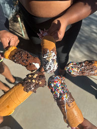 Sunlit overhead view of five hands holding chocolate-dipped churro cones topped with sprinkles, cookie crumbs and drizzled sauces over a concrete sidewalk