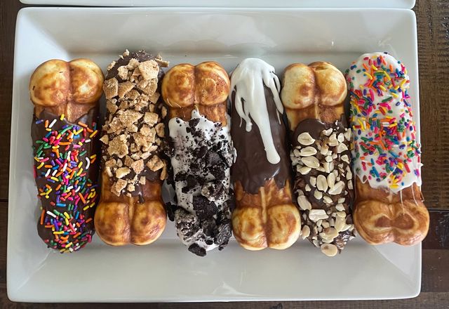 Six colorful chocolate-dipped bubble waffle sticks on a white rectangular plate over a wooden table, topped with rainbow sprinkles, crushed toffee, cookie crumbs, white chocolate drizzle, chopped peanuts, and white icing with sprinkles.
