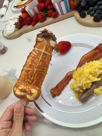 Playful brunch: chocolate-dipped waffle-on-a-stick held over a plate of scrambled eggs, bacon and sausage with a strawberry, with a wooden fruit board of strawberries, raspberries, blackberries and whipped cream in the background.