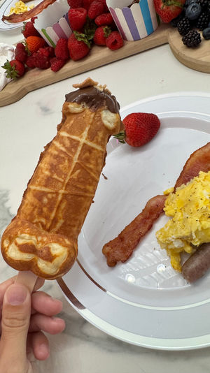 Playful brunch: chocolate-dipped waffle-on-a-stick held over a plate of scrambled eggs, bacon and sausage with a strawberry, with a wooden fruit board of strawberries, raspberries, blackberries and whipped cream in the background.