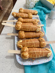 Novelty phallic-shaped golden waffle pops on wooden sticks arranged on a sprinkle-pattern platter on a kitchen countertop with a teal napkin — playful party dessert.