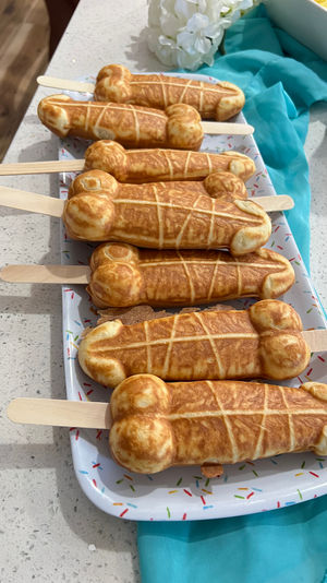 Tray of golden-brown novelty penis-shaped pancake pops on wooden sticks arranged on a sprinkle-patterned platter on a white kitchen countertop with turquoise cloth