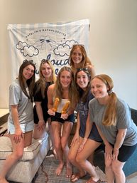 Six smiling women posing indoors in a living room group photo in front of a bachelorette party banner, bride-to-be holding two novelty-shaped pastry pops