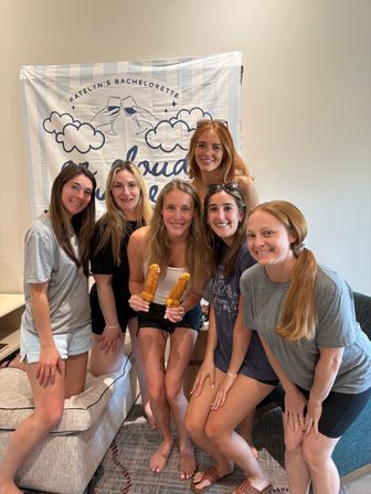 Six smiling women posing indoors in a living room group photo in front of a bachelorette party banner, bride-to-be holding two novelty-shaped pastry pops