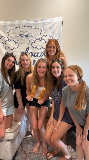 Six smiling women posing indoors in a living room group photo in front of a bachelorette party banner, bride-to-be holding two novelty-shaped pastry pops