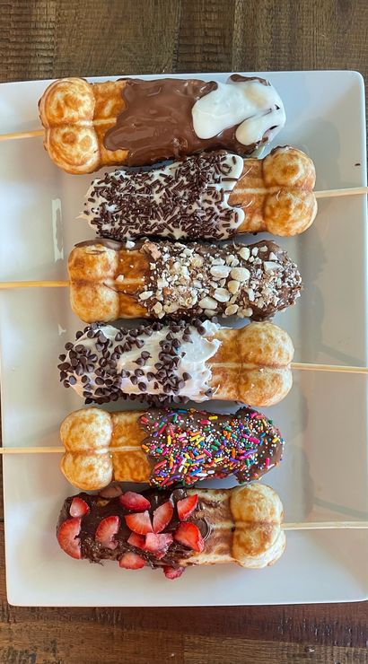 Six bubble-waffle sticks on a white rectangular plate — each waffle dipped in chocolate or white chocolate and topped with colorful sprinkles, mini chips, chopped nuts, chocolate sprinkles and fresh strawberries on a rustic wooden table.
