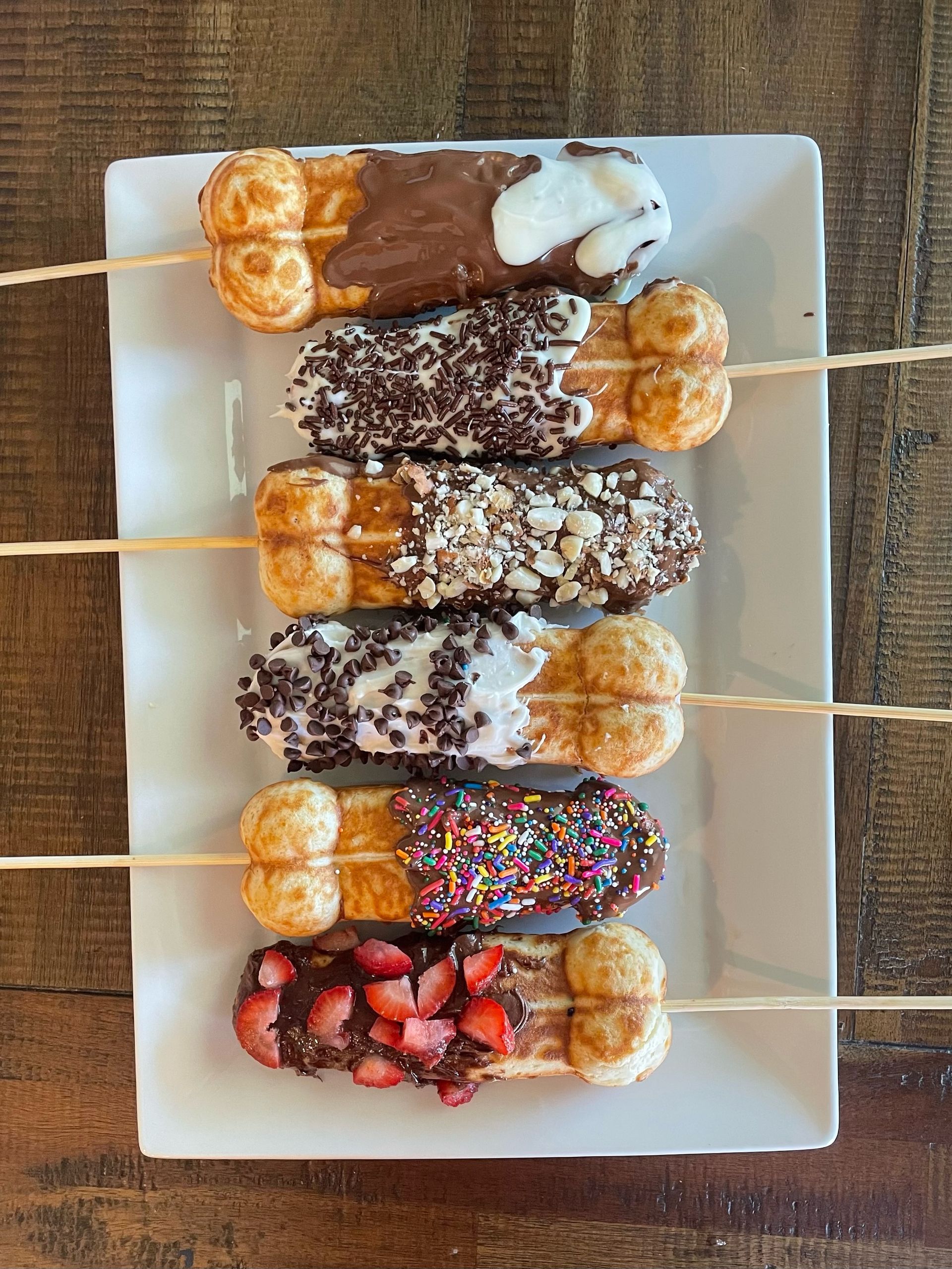 Six bubble-waffle sticks on a white rectangular plate — each waffle dipped in chocolate or white chocolate and topped with colorful sprinkles, mini chips, chopped nuts, chocolate sprinkles and fresh strawberries on a rustic wooden table.
