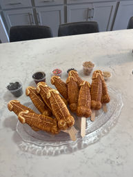Novelty penis-shaped waffle pops on wooden sticks arranged on a glass platter with small cups of sprinkles, chocolate sauce, crushed cookies and nuts on a white marble kitchen island