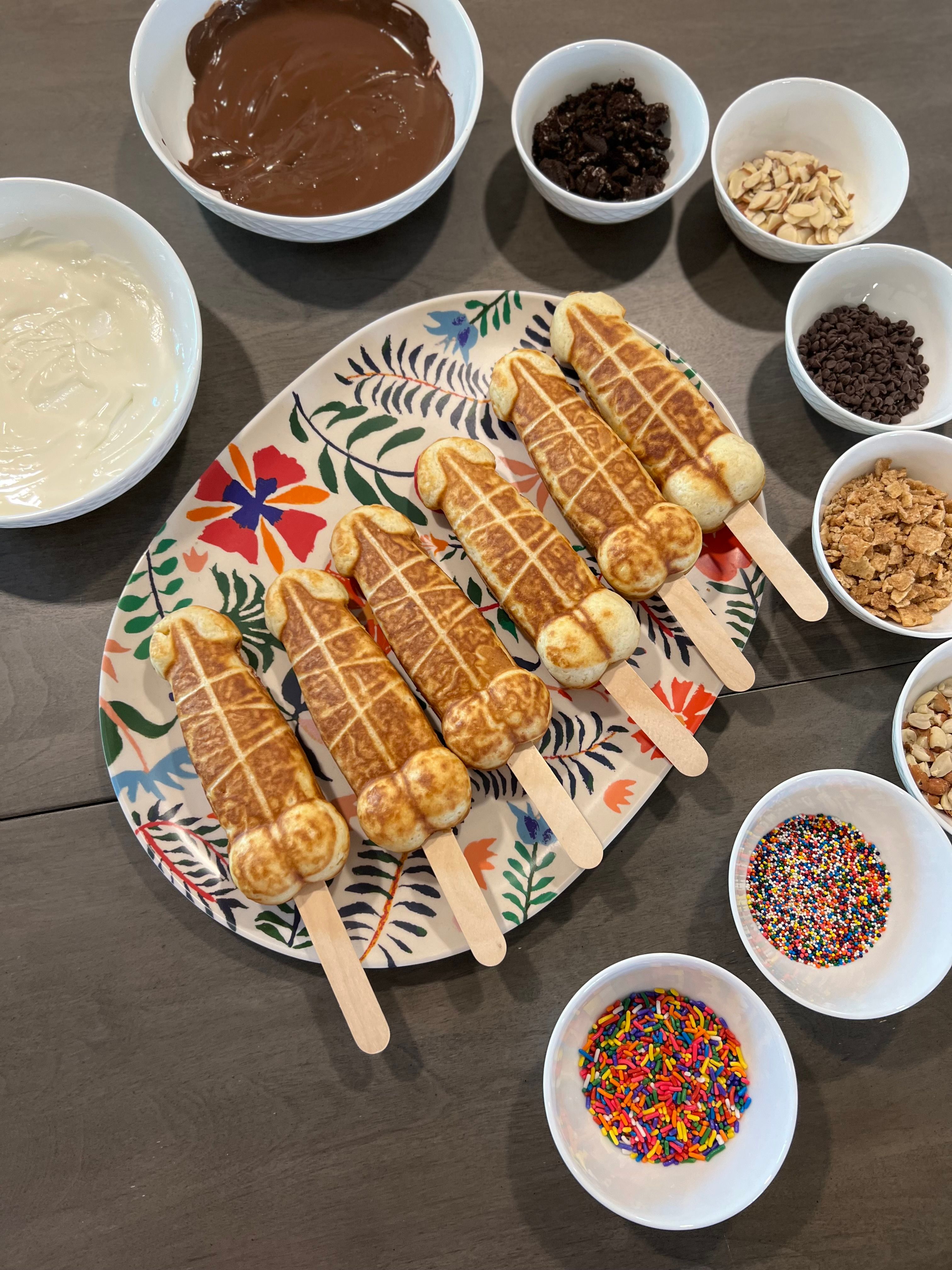 Six novelty waffle pops on wooden sticks arranged on a floral platter surrounded by bowls of melted milk and white chocolate, nuts, mini chips, crushed cookies, cereal and colorful sprinkles on a kitchen table — DIY dessert topping bar.