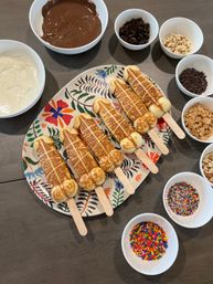 Six novelty waffle pops on wooden sticks arranged on a floral platter surrounded by bowls of melted milk and white chocolate, nuts, mini chips, crushed cookies, cereal and colorful sprinkles on a kitchen table — DIY dessert topping bar.