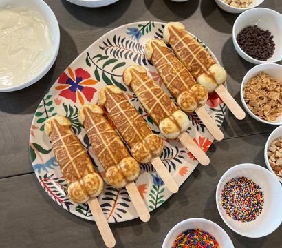 Six novelty waffle pops on wooden sticks arranged on a floral platter surrounded by bowls of melted milk and white chocolate, nuts, mini chips, crushed cookies, cereal and colorful sprinkles on a kitchen table — DIY dessert topping bar.