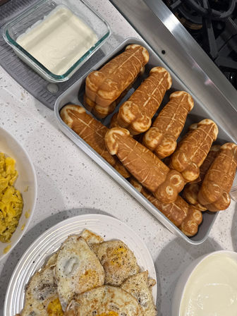 Home kitchen breakfast on a white quartz countertop: tray of golden tubular waffle sticks, plate of peppered fried eggs, bowl of scrambled eggs and bowls of yogurt or cream by the stove.