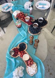 Backyard patio fruit charcuterie and dessert spread on a glass table with a turquoise fabric runner — wooden boards filled with strawberries, raspberries, blueberries, blackberries, mini marshmallows, whipped cream, chocolate dip, nuts, sprinkles and maple syrup, plated for a casual brunch.