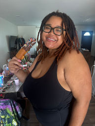 Smiling person with dreadlocks and glasses in a home living room holding a chocolate-coated treat on a stick topped with colorful sprinkles.