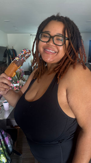 Smiling person with dreadlocks and glasses in a home living room holding a chocolate-coated treat on a stick topped with colorful sprinkles.