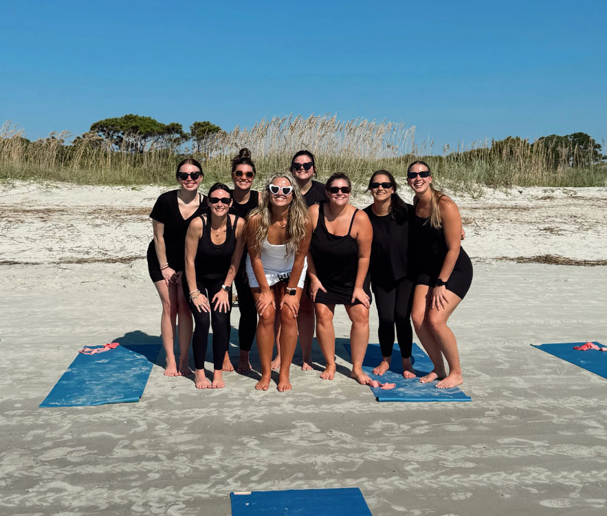 Nine women posing on blue yoga mats on a sunny sandy beach with coastal dune grass, mostly in black activewear and one in white with heart-shaped sunglasses, playful beach yoga group photo.