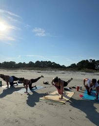 Sunny beach workout — a group doing plank variations with resistance bands on yoga mats along a sandy shoreline, blue sky and coastal trees in the background.