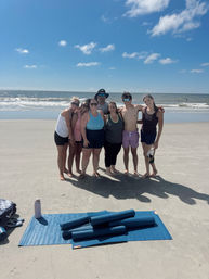 Seven people smiling and posing barefoot on a sunny sandy beach by the ocean, with blue sky, gentle waves, rolled-up yoga mats and a water bottle in the foreground.