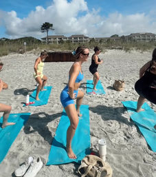 Group beach fitness class with women doing lunges on blue mats on a sunny sandy shore with dunes and beachfront buildings in the background.
