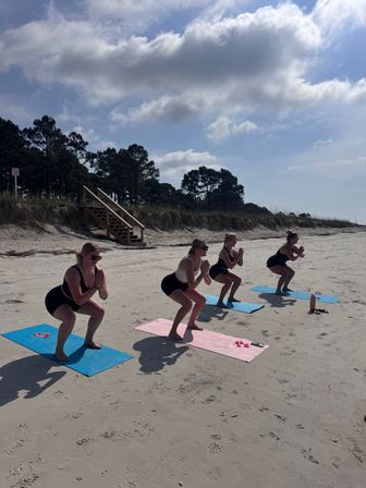 Four women in athletic wear doing squats on colorful yoga mats during a group beach workout on a sandy shore by dunes and wooden stairs under a partly cloudy sky