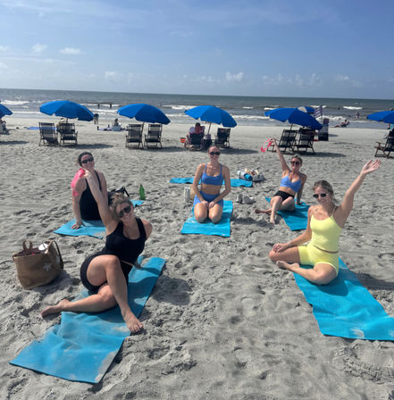 Five women in colorful activewear posing on blue yoga mats during a group beach yoga session on a sunny sandy shore with rows of blue umbrellas and gentle ocean waves