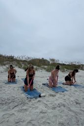 Group beach workout: four women kneeling on blue mats using pink resistance bands on sandy coastal dunes under a cloudy sky