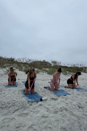 Group beach workout: four women kneeling on blue mats using pink resistance bands on sandy coastal dunes under a cloudy sky