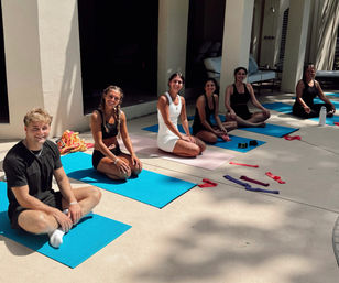 Six people seated on blue and pink yoga mats on a sunny poolside patio, smiling during an outdoor yoga/fitness session with resistance bands and a water bottle nearby.