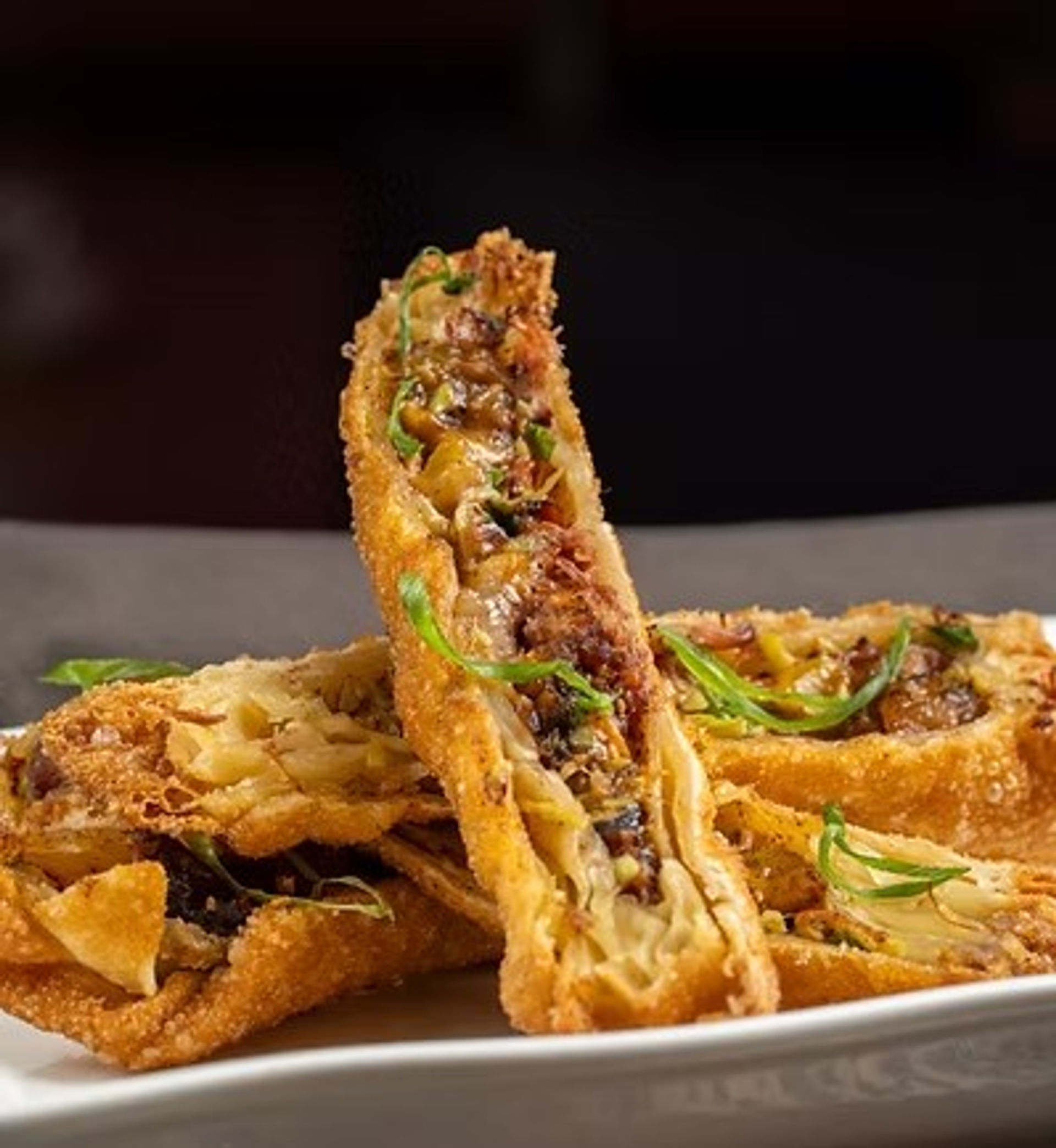 Crispy golden fried egg rolls cut open to reveal savory shredded meat and cabbage filling, garnished with sliced green onions on a white plate against a dark background.