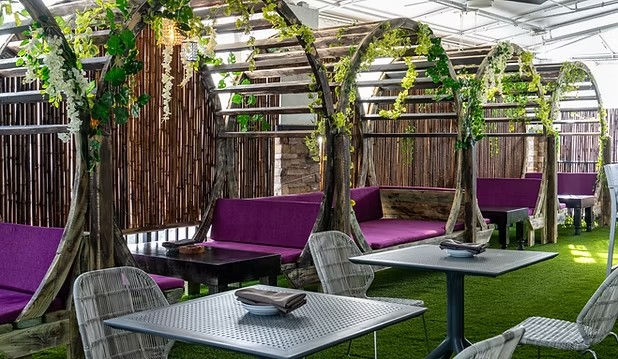 Covered outdoor cafe patio with curved wooden pergola booths draped in hanging greenery, purple cushioned benches, wicker chairs and metal tables on artificial turf.
