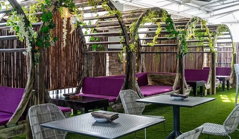 Covered outdoor cafe patio with curved wooden pergola booths draped in hanging greenery, purple cushioned benches, wicker chairs and metal tables on artificial turf.